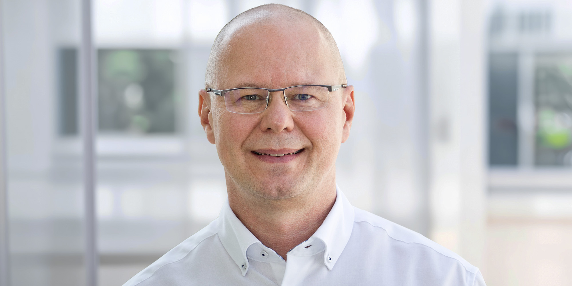 Jörg Klinge Jörg Klinge, lächelnd mit Brille und rasiertem Kopf, trägt ein weißes Hemd vor einem hellen, unscharfen Hintergrund.