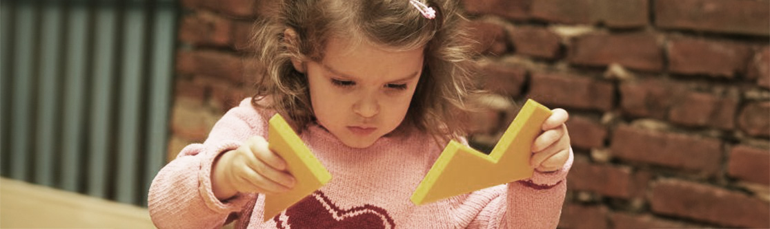 Entwicklung und Umsetzung Mädchen konzentriert sich beim Spielen mit zwei gelben Holzformen in einem Raum mit Backsteinwand.