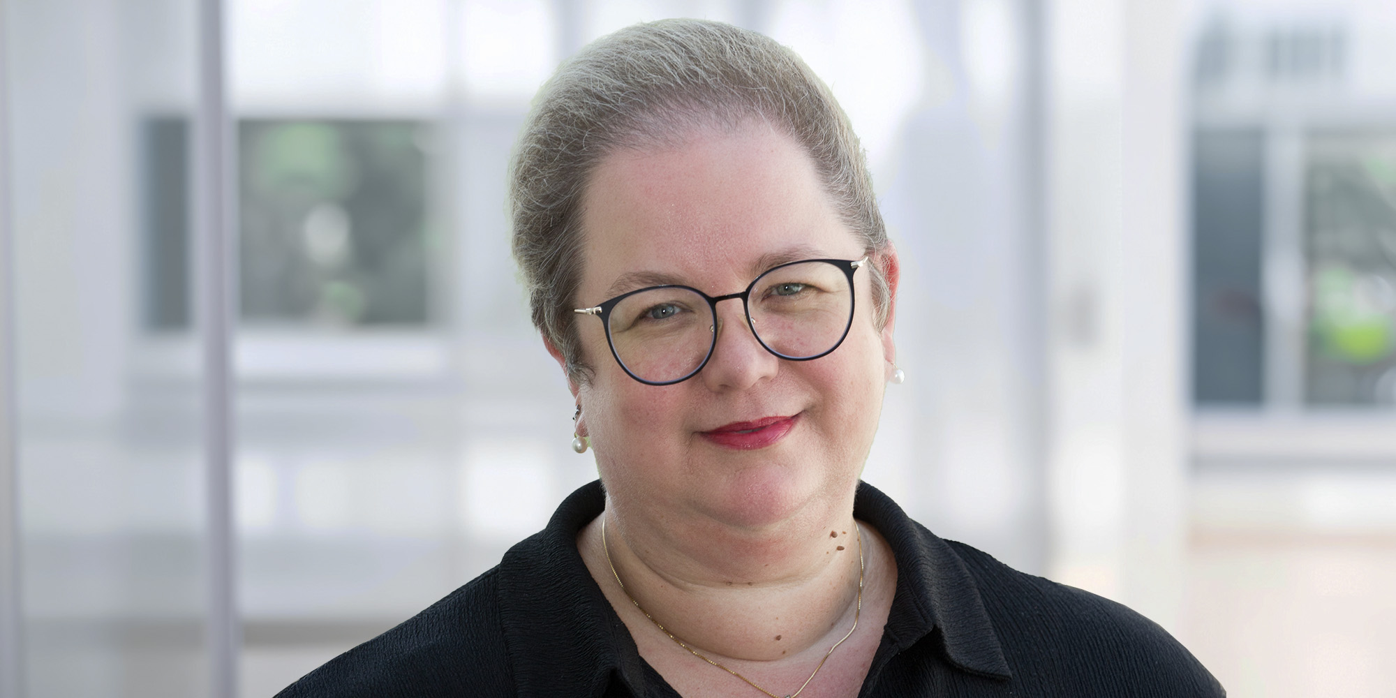 Claudia Fritz Claudia Fritz, lächelnd mit Brille und kurzem grauem Haar, trägt ein schwarzes Oberteil vor einem hellen, unscharfen Hintergrund.