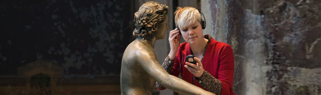 Entwicklung und kritische Reflektion Frau mit Kopfhörern betrachtet eine Bronzestatue in einem Museum und hört Informationen über ein Audioguide-Gerät.