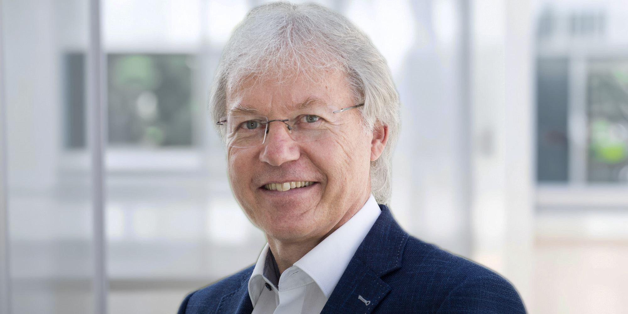 Michael Kraus Michael Kraus, lächelnd mit grauem Haar und Brille, trägt ein blaues Sakko und weißes Hemd vor einem hellen, unscharfen Hintergrund.