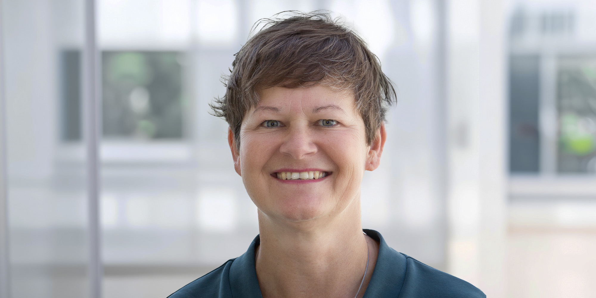 Nicole Schlabach Nicole Schlabach, lächelnd mit kurzem braunem Haar, trägt ein dunkelgrünes Oberteil vor einem hellen, unscharfen Hintergrund.