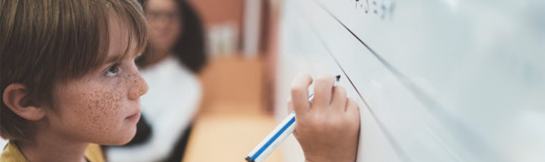 Förderung und Begleitung Junge schreibt mit einem Stift auf eine weiße Tafel in einem Klassenraum, im Hintergrund ist eine Lehrperson zu sehen.