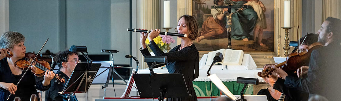 Mitwirkung im Auftrag oder für die Sparkassen-Finanzgruppe Musikerinnen und Musiker spielen klassische Musik in einer Kirche; eine Frau mit Querflöte steht im Mittelpunkt, umgeben von Streichinstrumenten, mit Altar und Gemälde im Hintergrund.