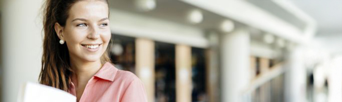 Information und Beratung Junge Frau mit langen Haaren und rosa Bluse hält Unterlagen in der Hand und lächelt, im Hintergrund ein modernes Gebäude.