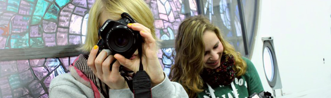 Mitwirkung bei Projekten Dritter Zwei junge Frauen bei der Arbeit mit einer Kamera, eine fotografiert, die andere lächelt, im Hintergrund bunte Glasfenster.