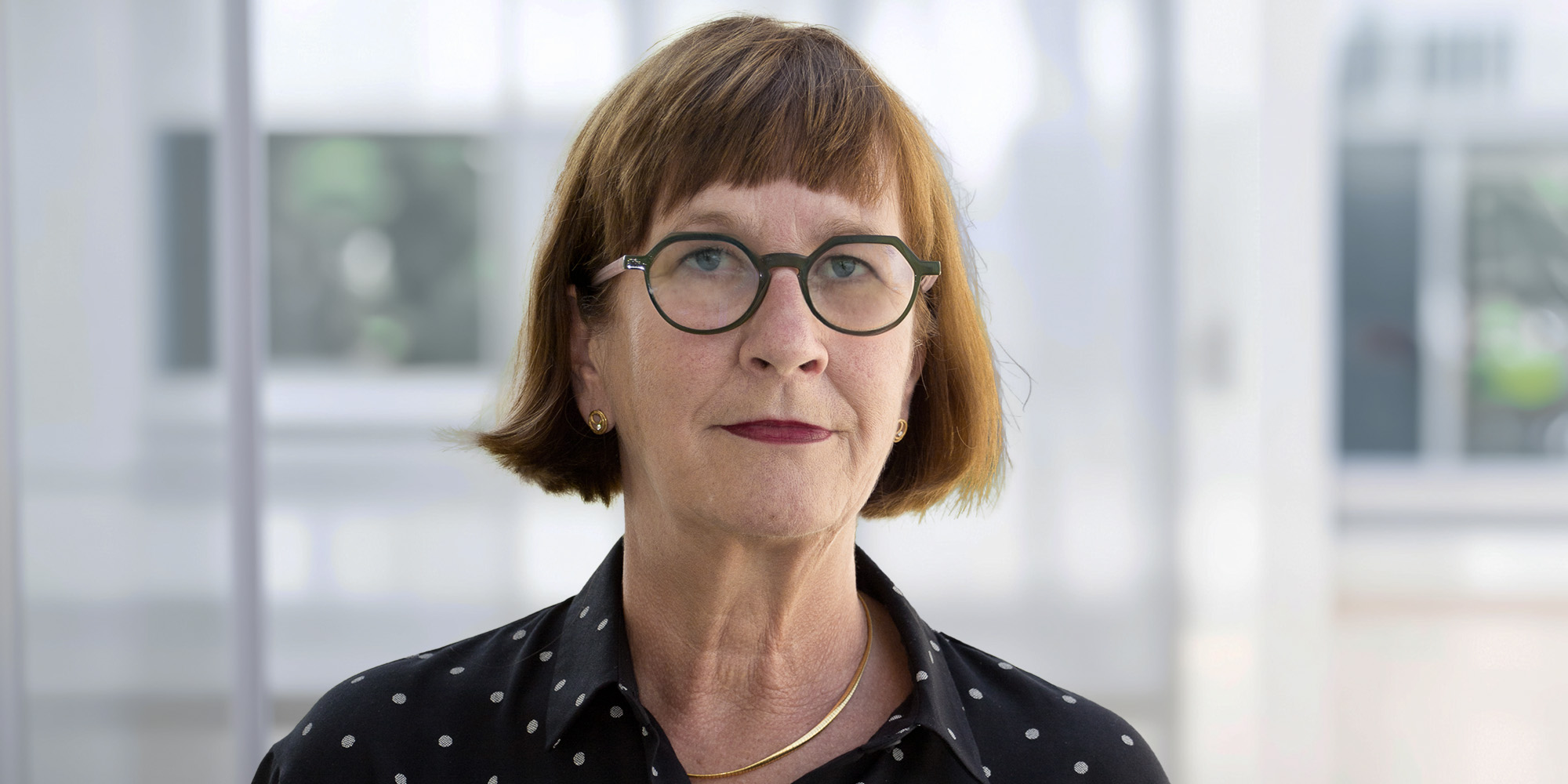 Bettina Riehl Bettina Riehl mit kurzem braunem Haar und Brille, trägt eine schwarze Bluse mit weißen Punkten vor einem hellen, unscharfen Hintergrund.